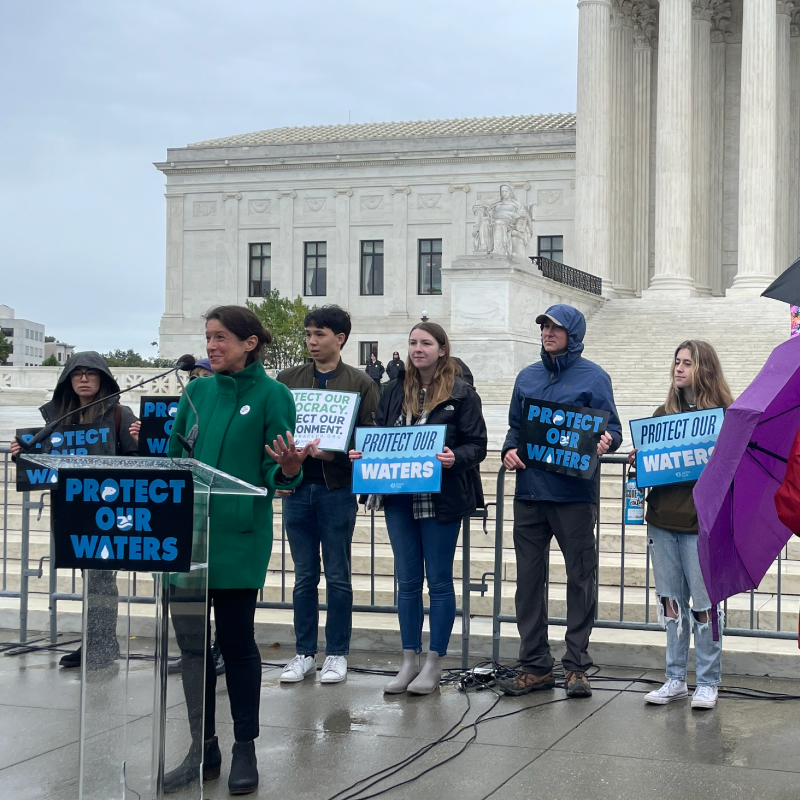 LCV's SVP of Government Affairs, Tiernan Sittenfeld, speaks at a podium in front of the Supreme Court building with people holding signs saying "Protect Our Waters"