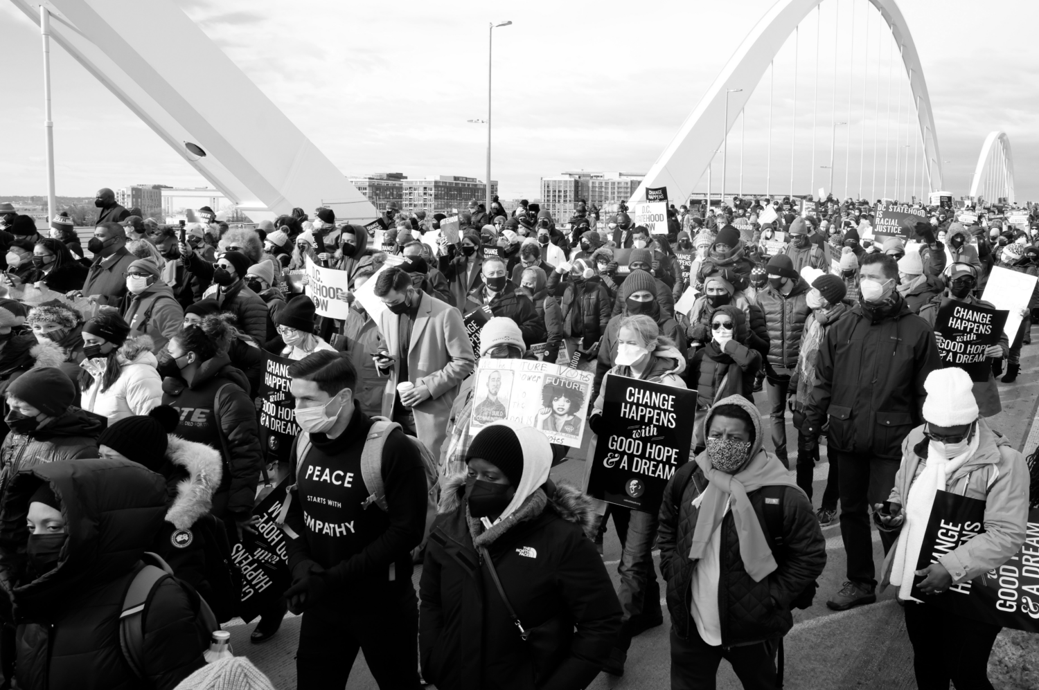 A black and white photo of crowd of people wearing masks and carrying signs reading "Change Happens with Good Hope and a Dream" march across a bridge.
