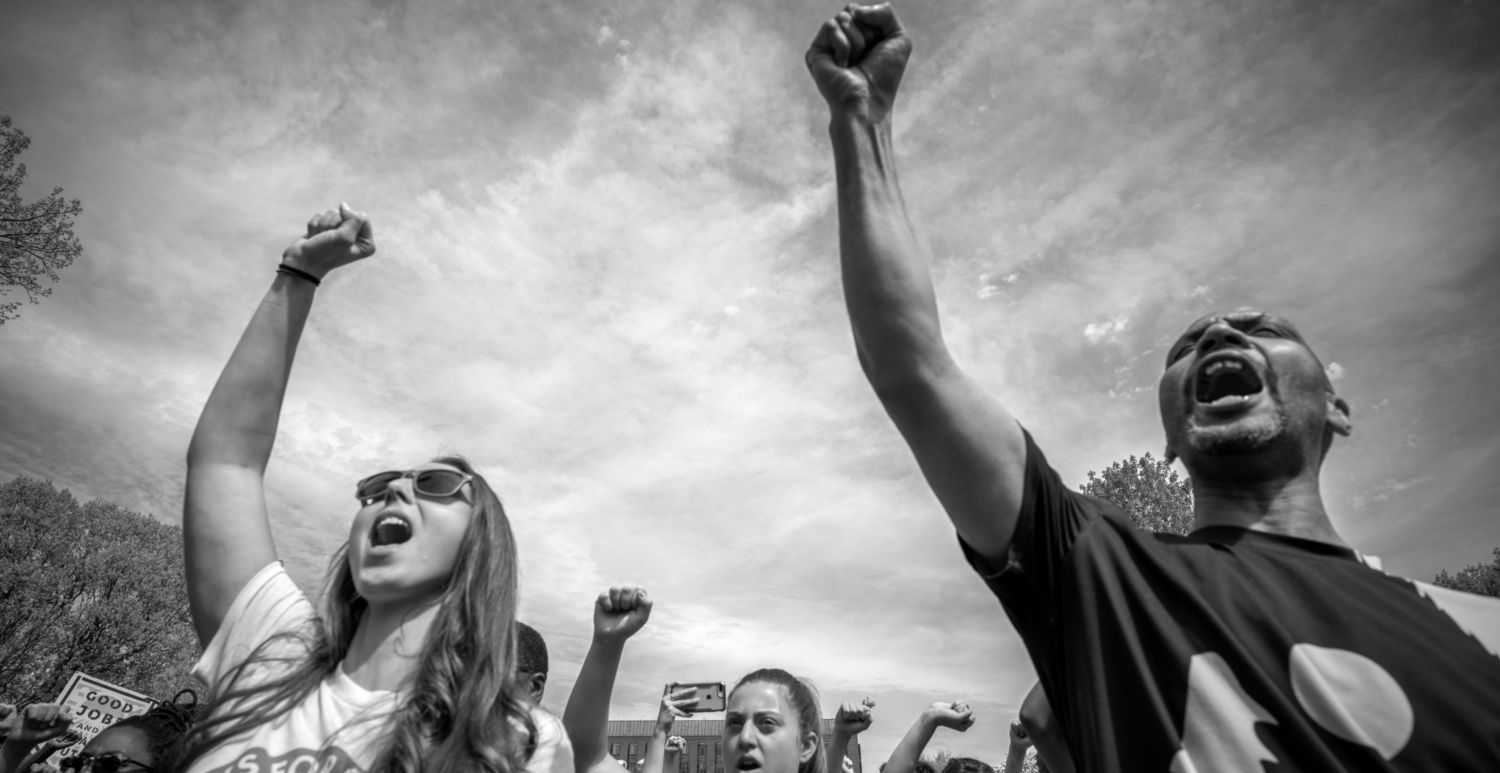 Black and white image of a man and a woman holding their fists in the air. Other fists are seen against the sky in the background.