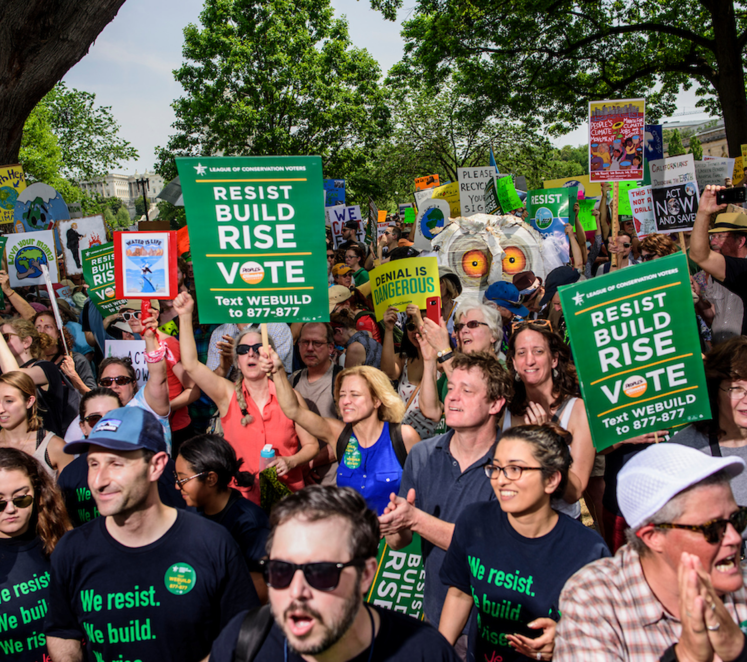 Image of crowd with people holding signs that read "Resist, Build, Rise, Vote" and "Denial is Dangerous".