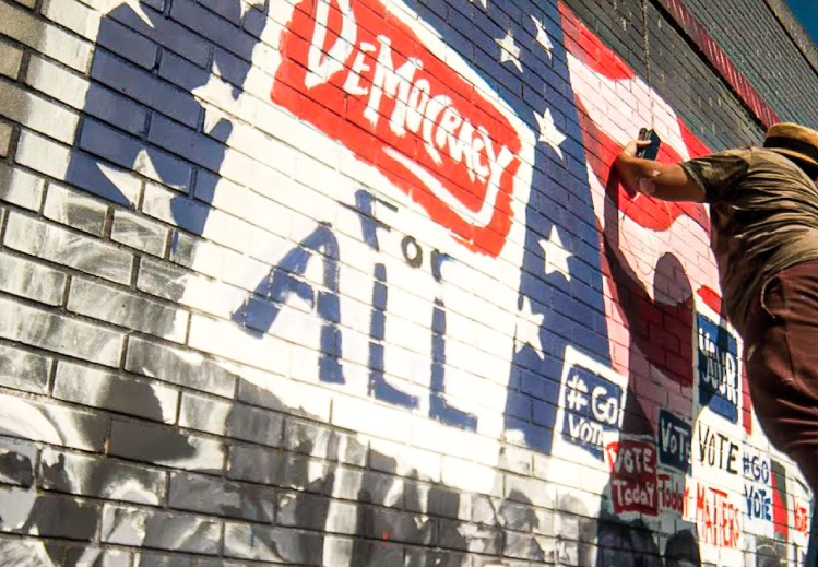 Painter on a ladder works on a mural that depicts protestors with signs reading "Democracy for All"