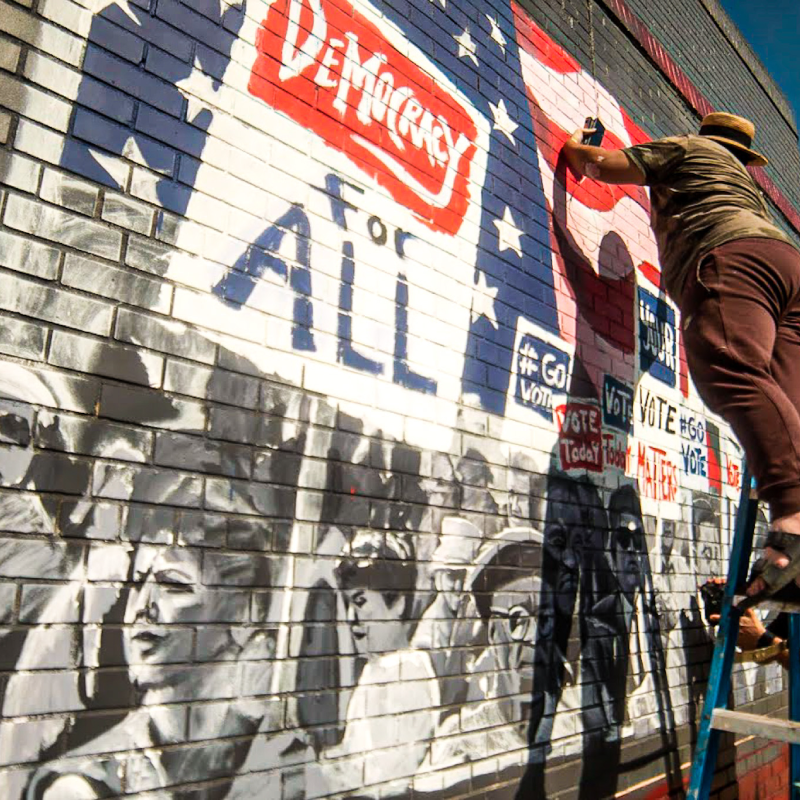 Painter on a ladder works on a mural that depicts protestors with signs reading "Democracy for All"