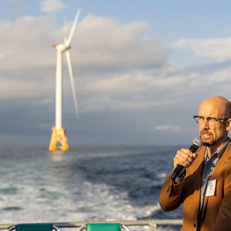 A clean energy activist speaks on a boat in front of an ocean wind turbine.