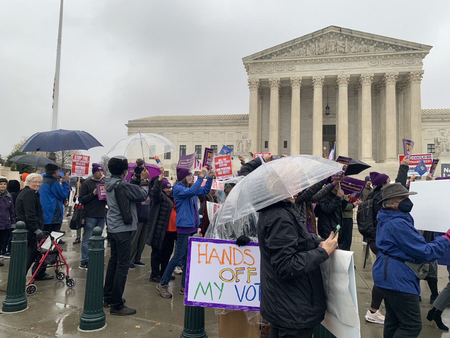 A crowd gathers outside the Supreme Court holding umbrellas and signs. One sign reads, "Hands off my vote"