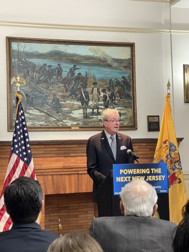 Governor Phil Murphy speaks at a podium that reads "Powering the next New Jersey". Behind him, the American flag, a fireplace, and a Revolutionary War painting.