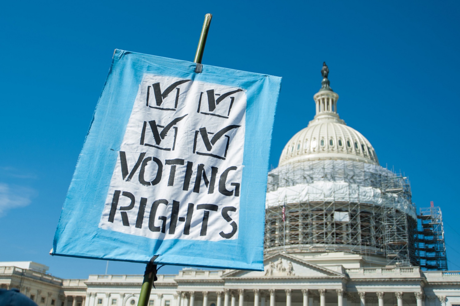 Foreground; A sign with four checkmarks that reads "Voting Rights" Background: The Capitol building under construction