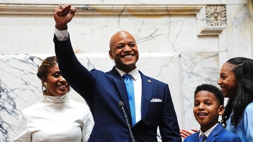 Governor Wes Moore smiles and holds his fist in the air. His children stand to his right and his wife stands behind him, smiling.