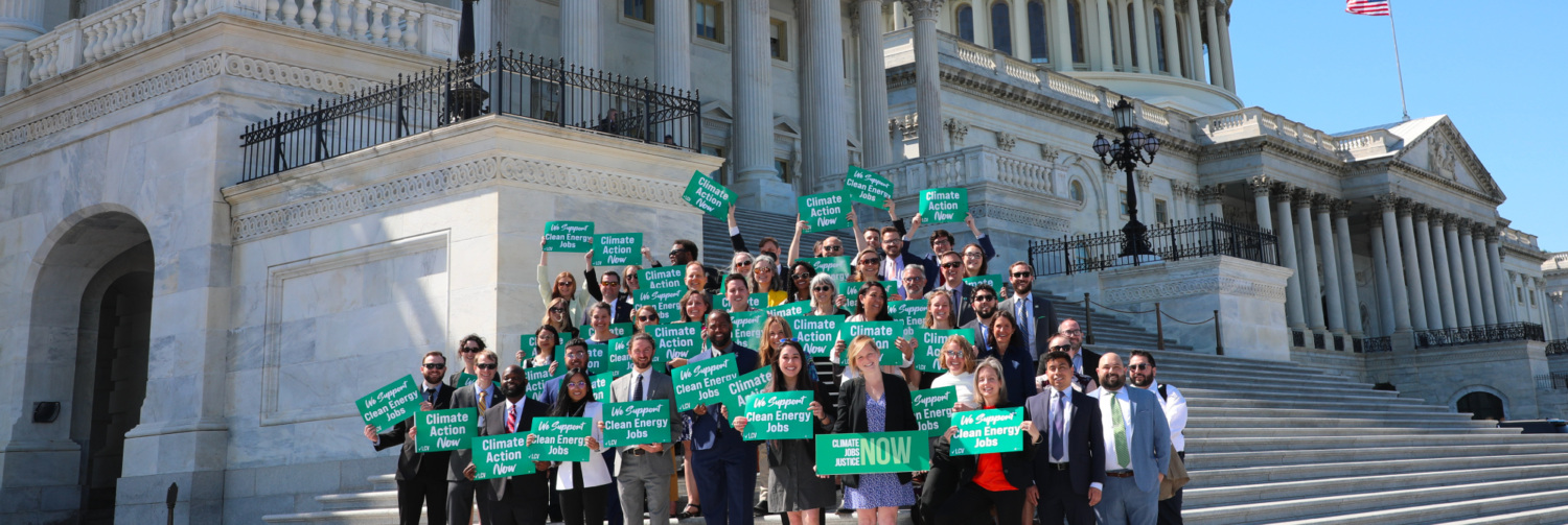 A large group of people from LCV and state league staff pose in front of Capitol building in DC with signs that read "Climate Action Now" and "We Support Clean Energy Jobs"
