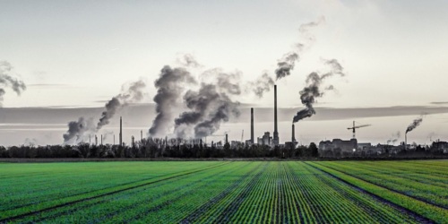 A large green farm field with an industrial skyline in the distance with billowing smog