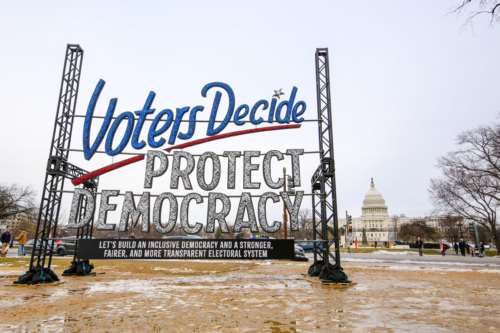 An art installation reads "Voters Decide, Protect our Democracy: Let's build an inclusive democracy and a stronger, fairer, and more transparent electoral system." The Capitol is seen in the background.