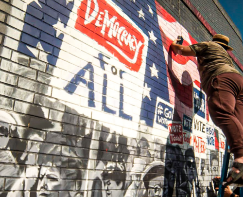 A painter works on a mural of protestors and a sign reading, "Democracy for all."