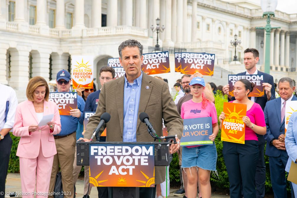 Representative John Sarbanes speaks at a podium in front of the Capitol and a group with signs reading "Freedom to Vote Act.”