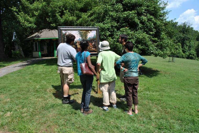 Four people look at a painting while standing on a grassy field dotted with green trees.