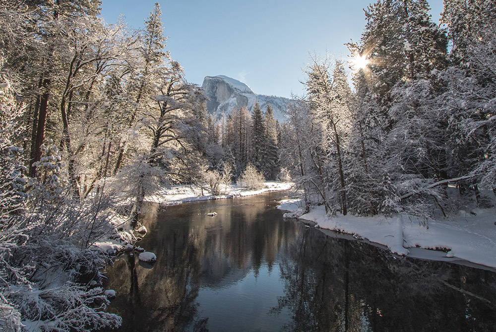 A snowy river landscape in Yosemite Valley with the Half Dome rock formation in the background.