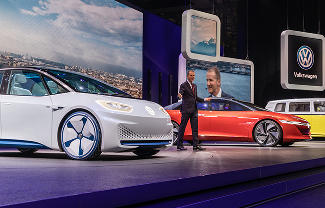 A Volkswagon executive giving a talk amongst electric vehicles on stage.