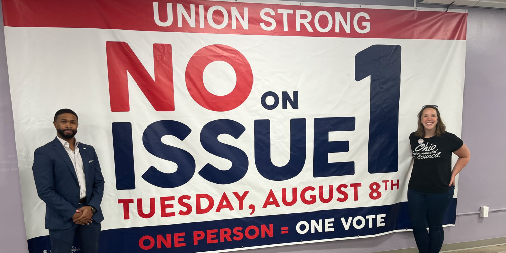 Two people stand on either side of a large sign. The sign reads "Union Strong: No on Issue 1. One Person = One Vote."
