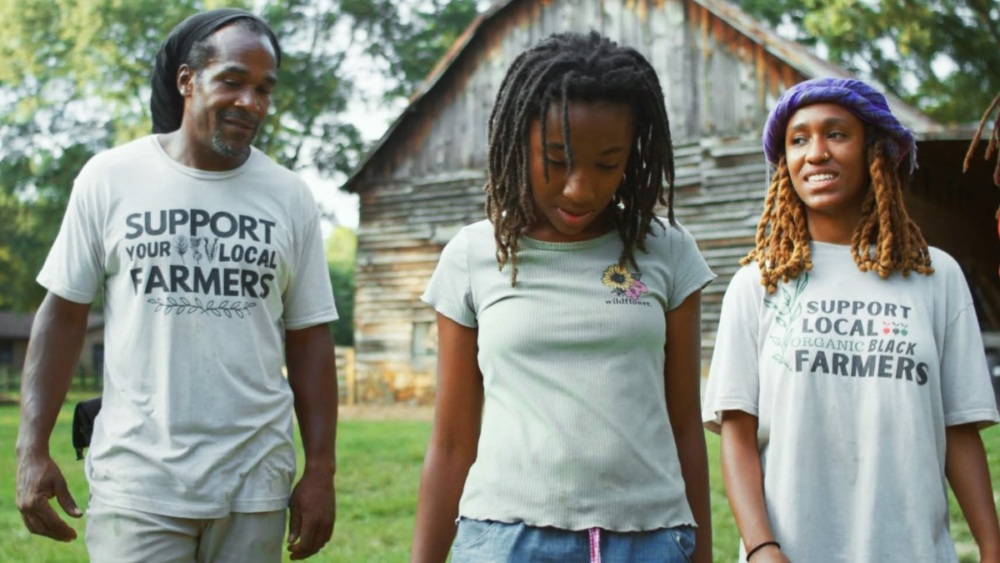 A man and two girls in front of a barn, all wearing green shirts with one reading “Support Your Local Farmers” and another “Support Local Organic Black Farmers.”