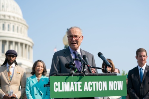 Senator Schumer speaks at a podium with a "Climate Action Now" sign and the Capitol dome in the background.