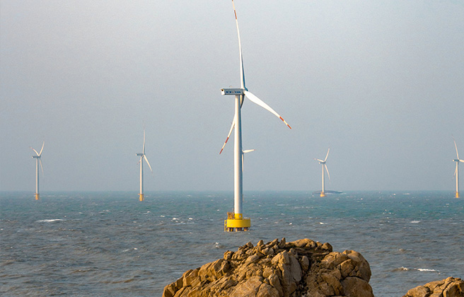 White wind turbines in the ocean with large rocks in the foreground.