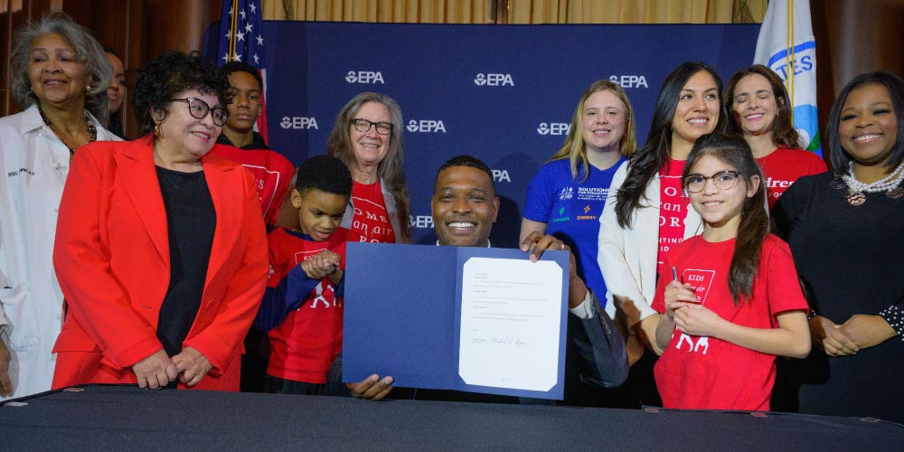 EPA Administrator Michael S. Regan poses at signing of new soot pollution standards, surrounded by several smiling women and children