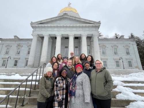 Group of 14 people bundled in coats, posing on the snowy steps of the Vermont state capitol building in Montpelier.