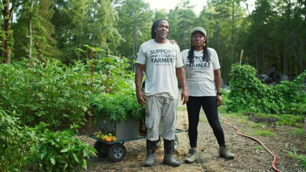 A man and woman wearing shirts reading "Support Your Local Farmers" stand together amid a field of different vibrant green crops.