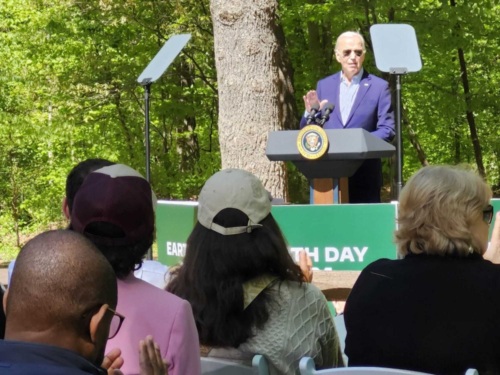 Photo taken from behind a row of audience members applauding President Biden as he speaks at a podium with forest trees in the background.