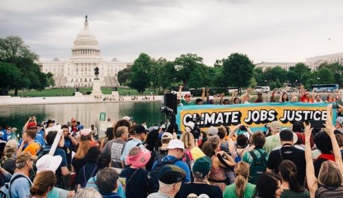 A crowd of Earth Day marchers with a sign reading "Climate, Jobs, Justice" and the Capitol building visible in the background.