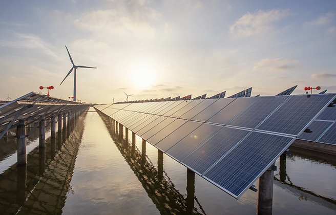 View down a row of solar panels with a wind turbine in the background.