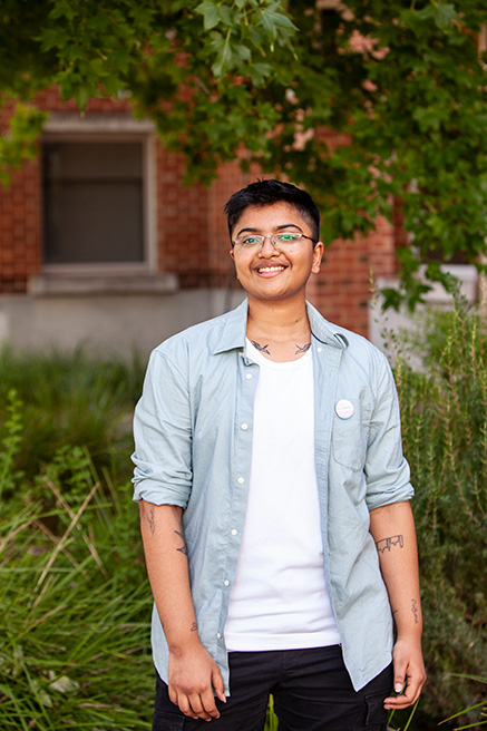 Krishna stands outdoors in front of a building and some greenery.