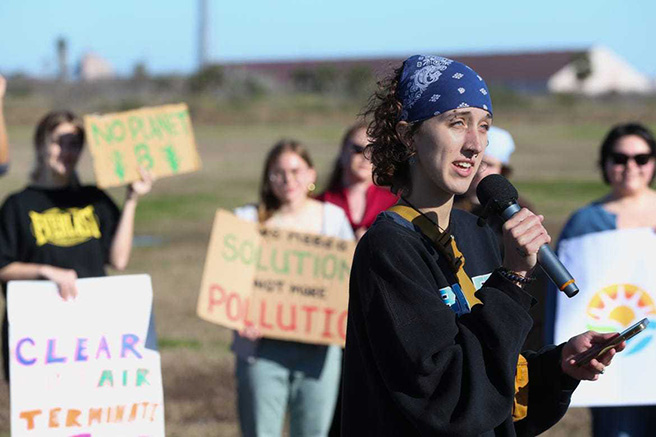 Maggie stands outdoors in front of a group of activists holding signs and speaks into a microphone.