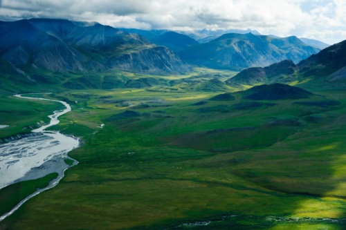 A river curves through a lush green landscape with a mountain range rising above it, topped by puffy clouds.