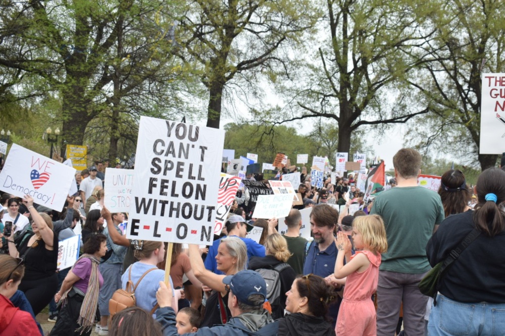 A crowd of protesters with signs in the foreground reading "We [heart] Democracy!" and "You can't spell Felon without Elon."