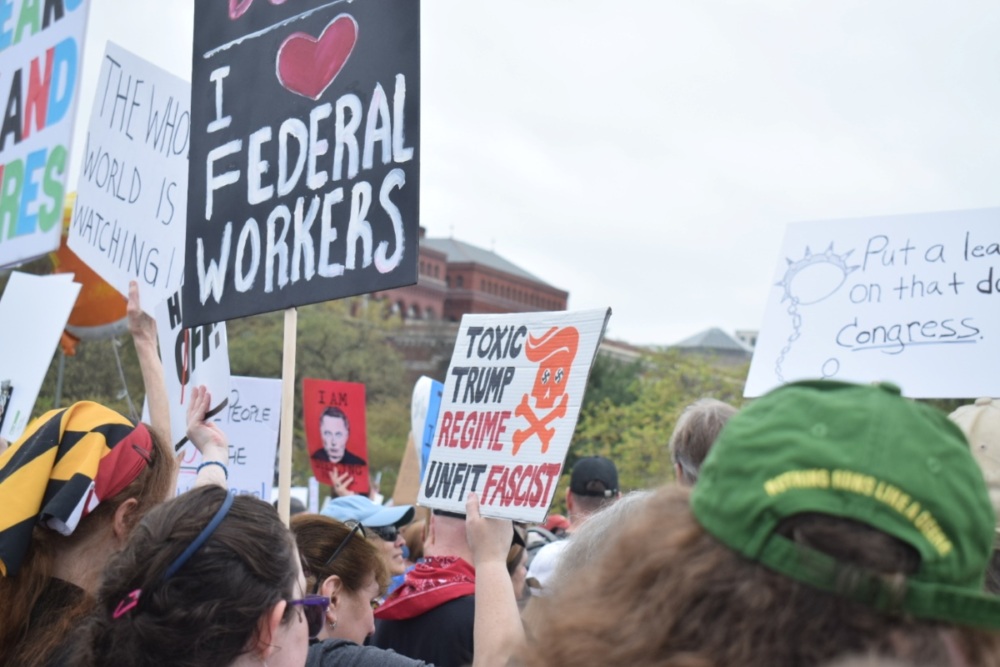 Protest signs including one reading "I [heart] federal workers," and another "The whole world is watching."