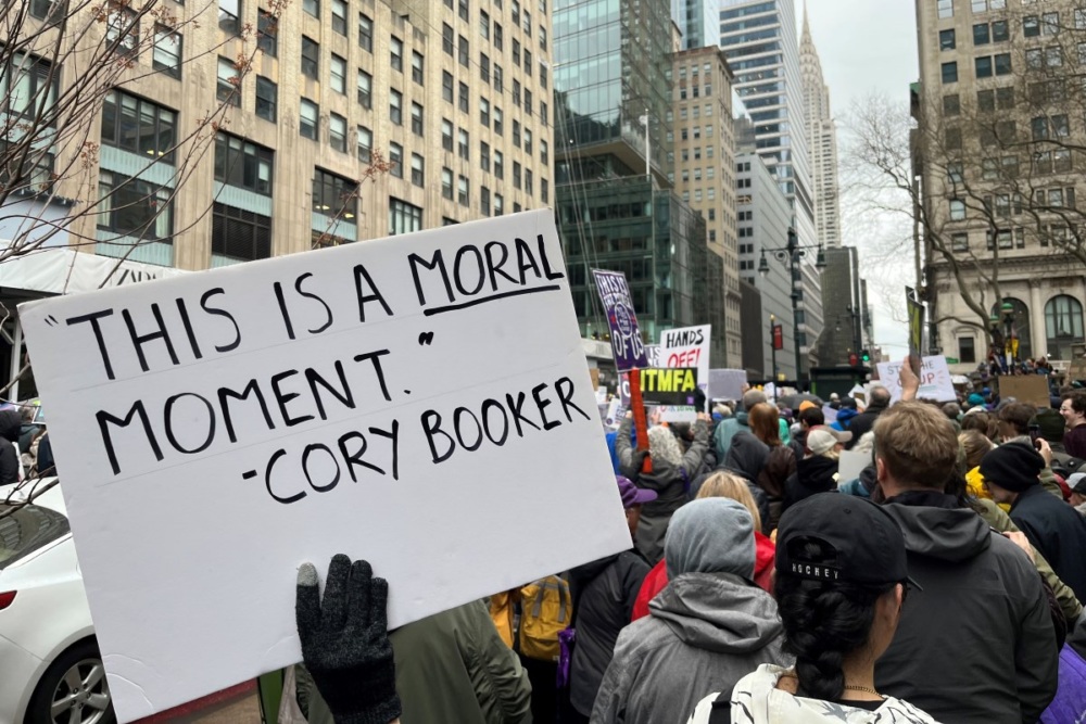 Protesters march in New York City, and a sign in the foreground reading "This is a moral moment - Cory Booker."