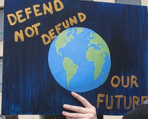 A protest sign with a picture of the Earth, reading "Defend not defund our future."