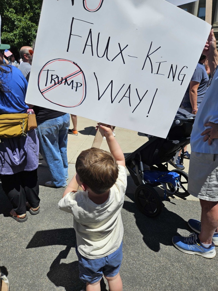 A child is pictured from behind holding a sign that reads "No Faux-King Way!"