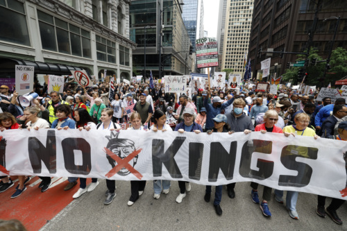 Protestors carry a banner that reads “No Kings” and features an image of Trump wearing a crown with an X covering his face.