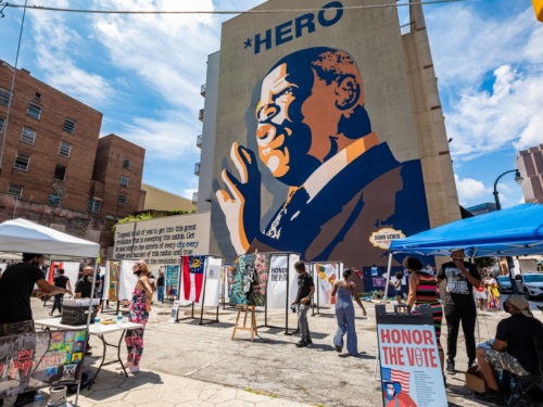 A huge mural on the side of a building that portrays John Lewis towers in the background as people visit tables and explore other art installations at a get-out-the-vote event.