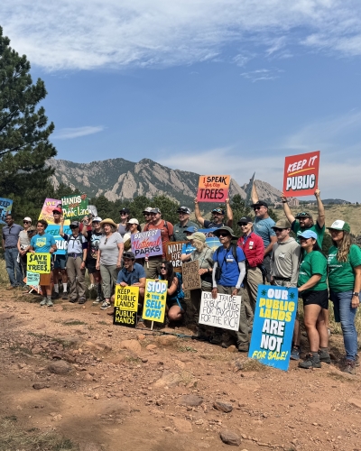 A group poses by a trail holding signs with messages including "Public Lands are not for sale" and "Stop the sell-off"