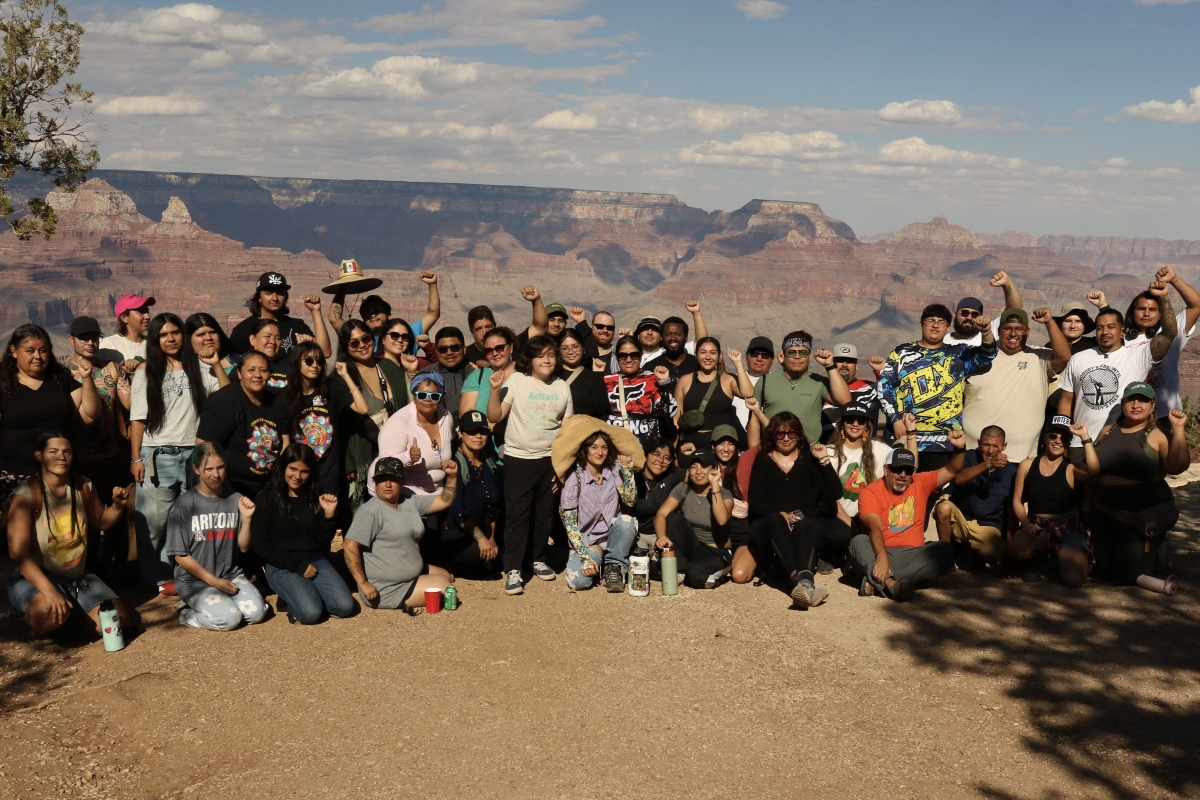 A large smiling group poses on a trail with the cliffs of the Grand Canyon in the background.