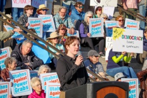 A person with a microphone speaks as a group listens, many holding blue signs reading "Utahans Against Power Grabs" and other pro-democracy posters.