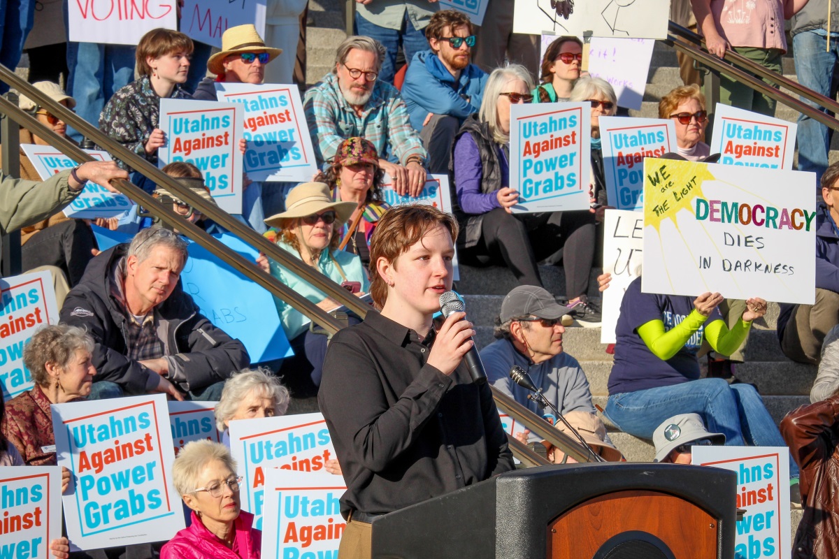 A person with a microphone speaks as a group listens, many holding blue signs reading "Utahans Against Power Grabs" and other pro-democracy posters.