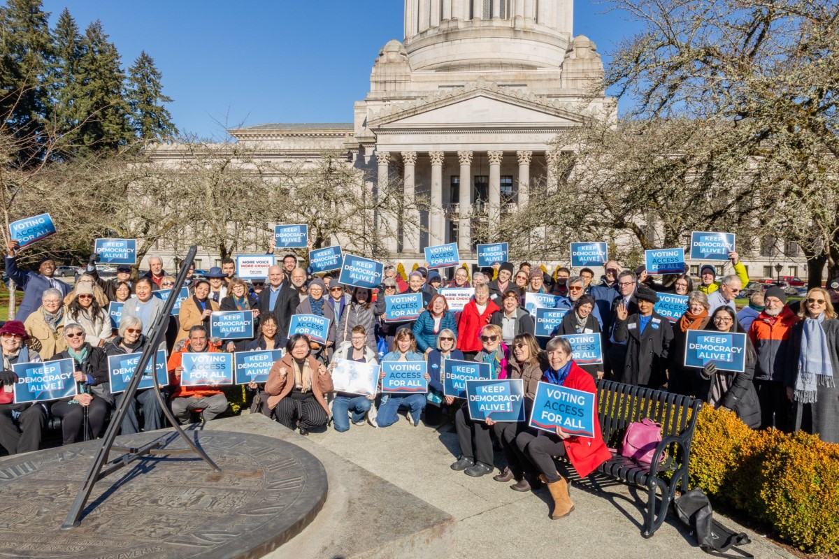 A group sitting in front of the Washington state capitol, holding blue signs reading "Keep Democracy Alive!"