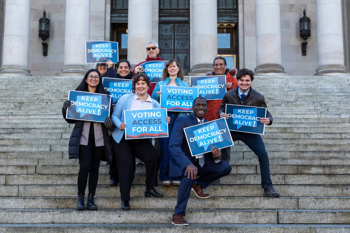 A group of nine people on the steps of the Washington state capitol, holding blue signs reading "Keep Democracy Alive!"