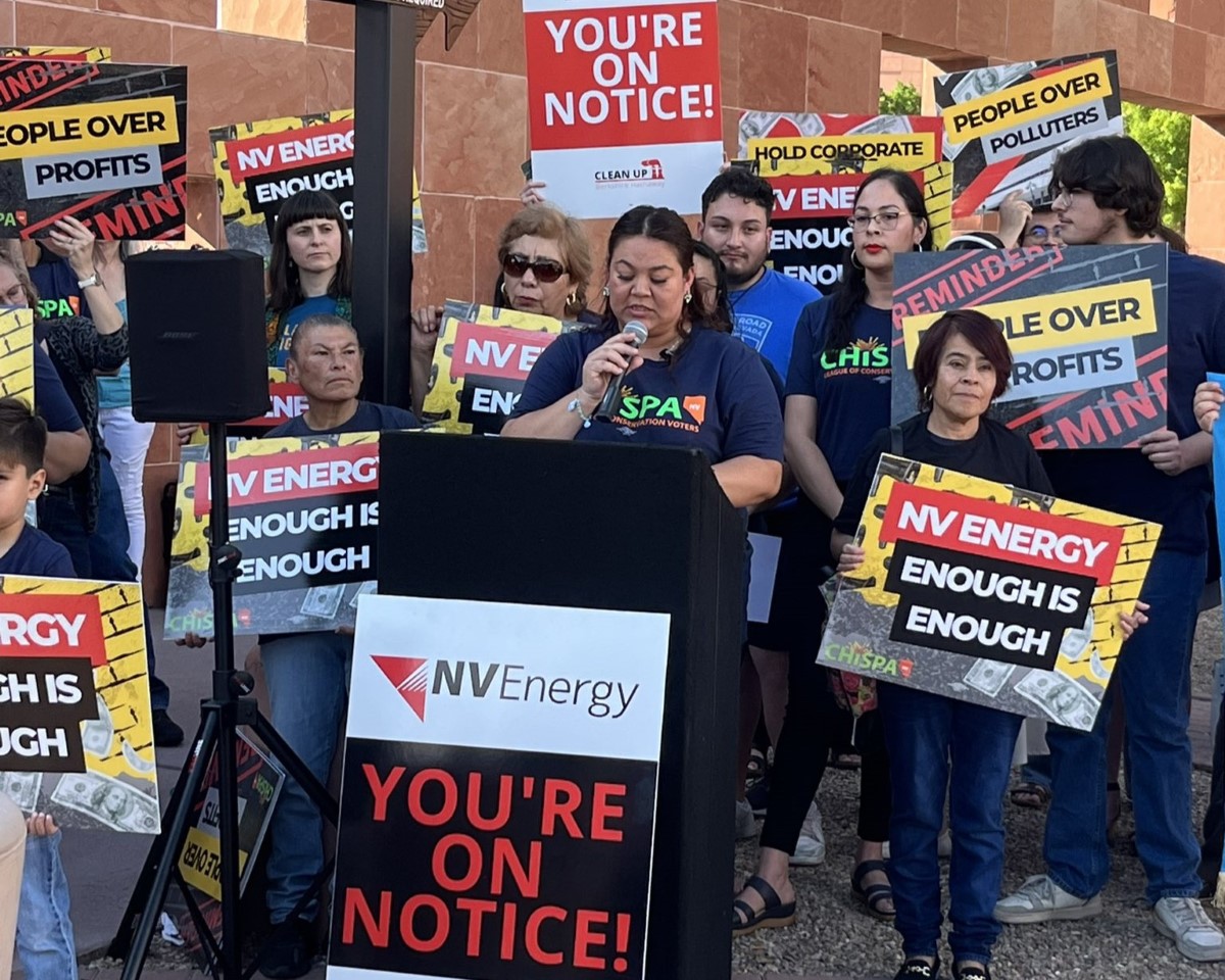 A woman speaks at a podium. Behind her is a group of people holding signs that read, "NV Energy: Enough is enough."