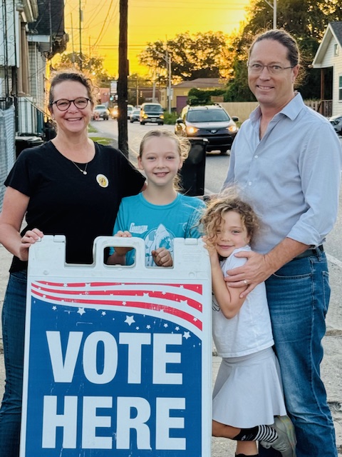 A man and woman pose with their two young daughters next to a "Vote Here" sign.