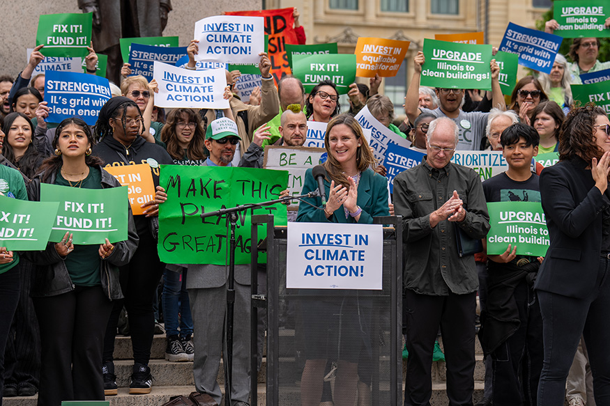 A woman stands at a podium surrounded by people holding signs with messages supporting climate action.