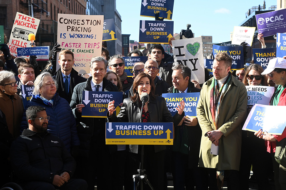 Governor Kathy Hochul stands at a podium surrounded by supporters holding signs supporting congestion pricing.