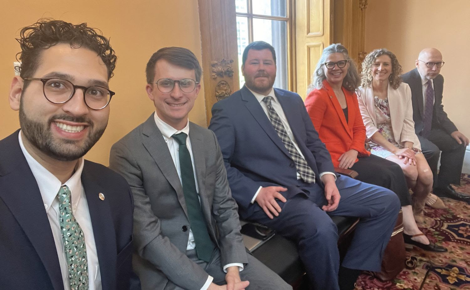 A group of 5 people sit on a bench in front of a window.